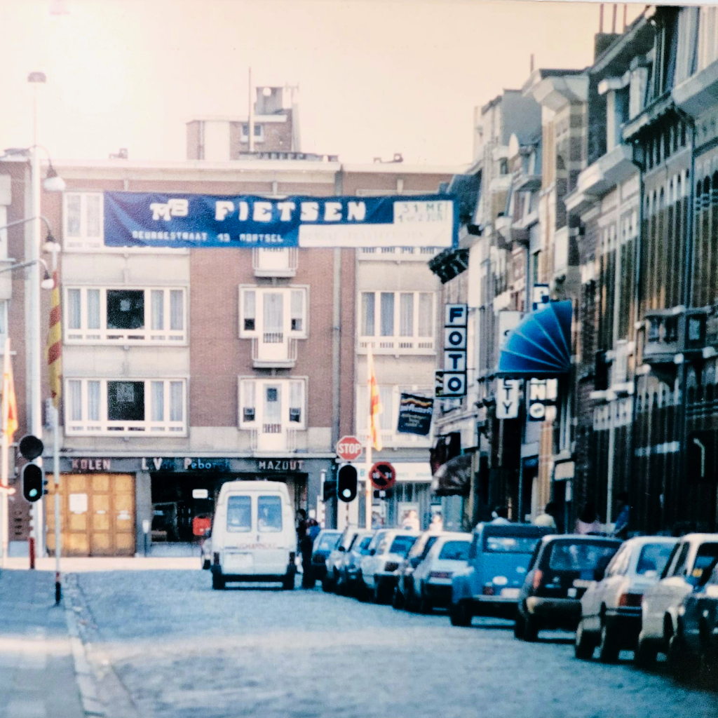 Hier ziet u een foto van de Deurnestraat in Mortsel van in de jaren tachtig. Er hang een banner met daarop de aankondiging van de Deurnestraat feesten, een rommelmarkt in Mortsel